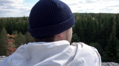 Bulutlu bir gökyüzü ve ağaçlar tepeler seyir yüksek dağın tepesinde oturan yalnız adam. Bir kayaya oturan şapka ve beyaz ceketli adam güzel orman arayan bir arka görünümü.