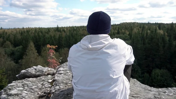 Hombre solo sentado en una cima de alta montaña mirando un cielo nublado y picos de árboles. Vista trasera de un hombre con sombrero y chaqueta blanca sentado en una roca, mirando el hermoso bosque . — Foto de Stock
