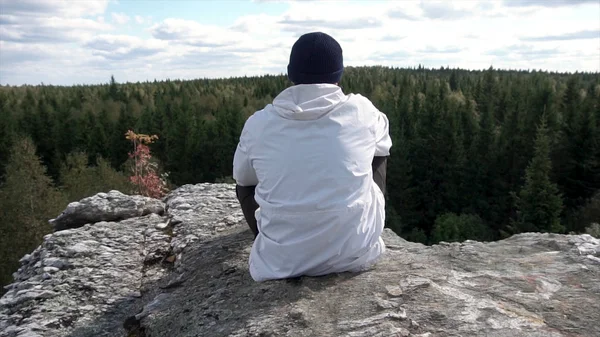 Hombre solo sentado en una cima de alta montaña mirando un cielo nublado y picos de árboles. Vista trasera de un hombre con sombrero y chaqueta blanca sentado en una roca, mirando el hermoso bosque . — Foto de Stock