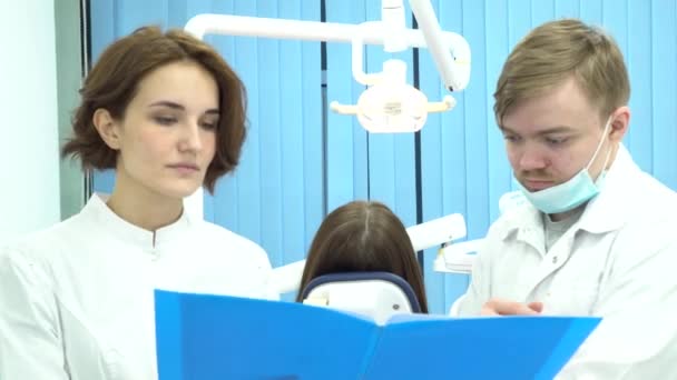Kvinnliga och manliga tandläkare hålla en mapp med dokument som diskuterar behandling framför patienten i stol. Läkare läsa Mappanteckningar i medicinsk skåpet. — Stockvideo