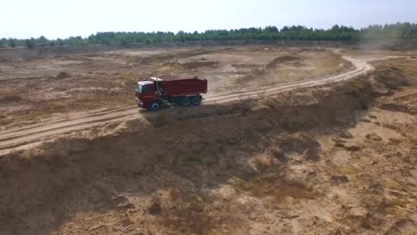 Αεροφωτογραφία του άδειο κόκκινο φορτηγό απορρίψεων για άμμο παράδοση κινείται στην άμμο πίστα με το πεδίο και τον ουρανό στο παρασκήνιο. Σκηνή. Εκτός δρόμου φορτηγό. — Αρχείο Βίντεο