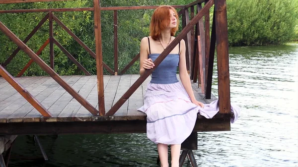 Mujer joven, delgada, pelirroja, sentada en una cubierta junto al agua, mirando a la distancia. Chica reflexiva brillante sentado en el muelle en falda larga cerca del lago . —  Fotos de Stock