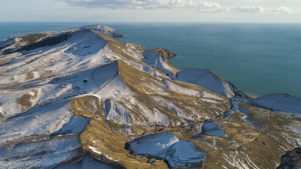 Luchtfoto van besneeuwde rotsen, blauwe zee, op bewolkte hemelachtergrond. Schot. Besneeuwde heuvels in IJsland door de zee in een winterdag. — Stockfoto