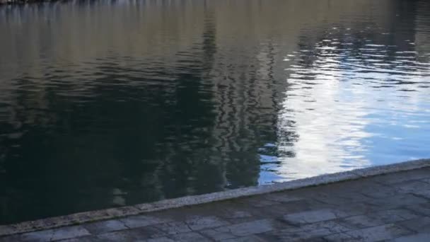 Close up for grey sidewalk near a river with a fountain. Stock. Asphalted footpath near dark water of the river with the reflection of blue, cloudy sky. — Stock Video
