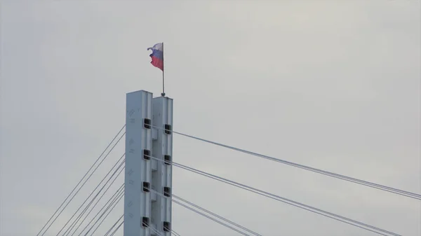 Bandeira da Federação Russa balançando ao vento no topo da ponte. Estoque. Bandeira russa no topo de uma ponte em cinza, fundo céu nublado . — Fotografia de Stock