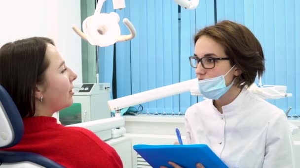 Vrouwelijk tandarts in tandheelkundige kantoor bedrijf van map en praat met de vrouw patiënt. Tandheelkundige specialist in de kliniek kabinet gevraagd jonge vrouwelijke patiënt over haar tanden. — Stockvideo
