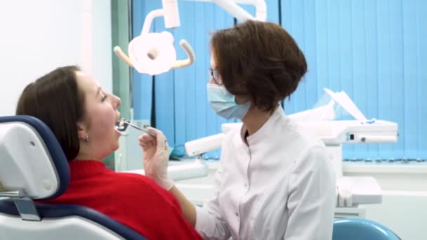 Κορίτσι οδοντίατρος φινίρισμα να εξετάσει τη στοματική κοιλότητα του ασθενούς γυναίκα κάθεται στη καρέκλα του οδοντιάτρου, οδοντιατρική φροντίδα έννοια. Νέος οδοντίατρος στο labcoat και μια μάσκα κατά την διαδικασία επεξεργασίας. — Αρχείο Βίντεο