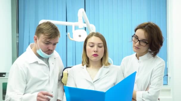 Grupo de pasantes o médicos jóvenes con batas de laboratorio blancas que miran la carpeta azul, la medicina y el concepto de atención médica. Tres dentistas en el gabinete discutiendo el diagnóstico decepcionante, se ven tristes . — Vídeo de stock