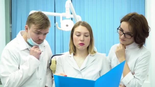 Group of interns or young doctors in white lab coats looking at blue folder, medicine and health care concept. Three dentists in the cabinet discussing disappointing diagnosis, lookig sad. — Stock Video