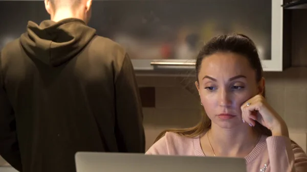 Cute woman working on the laptop in the kitchen while her boyfriend standing making her coffe or tea. Man putting a mug on the table for a woman with laptop, hugging her and kissing in forehead. — Stock Photo, Image