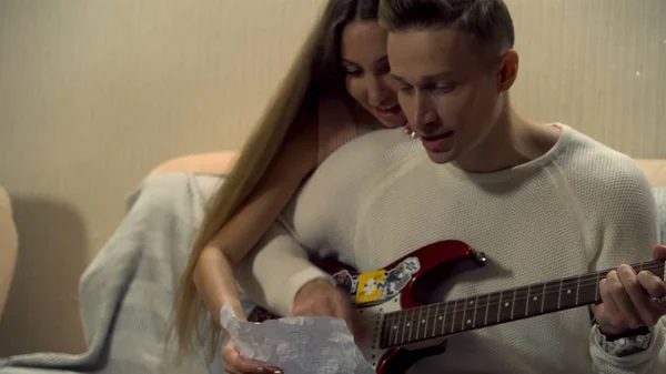Man sitting on a couch, playing guitar with a hug from his girlfriend. Young man is playing the electric guitar, young pretty woman is sitting behind him, hugging him, singing.