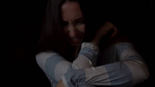 Portrait of a sad, young woman hugging her knees and crying on dark background, loneliness concept. Depressed female sitting alone and crying against the dark wall.