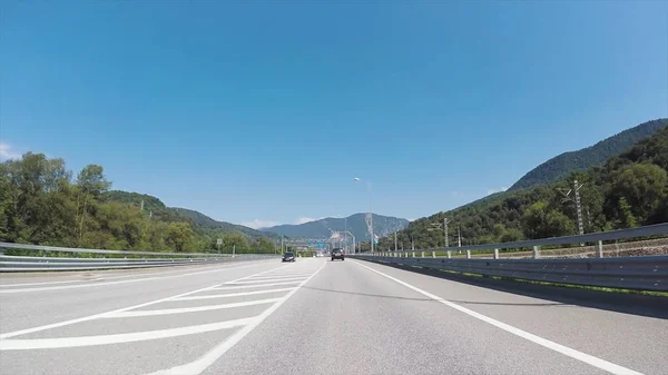 Vue de la route par la fenêtre de la voiture se déplaçant sur l'autoroute parmi les arbres par une journée ensoleillée d'été. Scène. Véhicule roulant sur la route contre le ciel nuageux et bleu et les montagnes boisées . — Photo