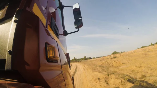 Side view of red truck cab moving on dusty unpaved rural road along green trees ahead. Scene. Huge lorry riding on country, dusty road on cloudy sky background, view from a wheel. — Stock Photo, Image