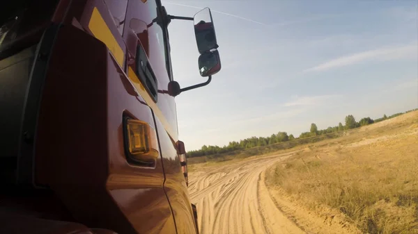 Seitenansicht eines roten LKW-Führerhauses, das sich auf einer staubigen, unbefestigten Landstraße entlang grüner Bäume voran bewegt. Szene. Riesenlaster auf dem Land, staubige Straße vor wolkenverhangenem Himmel, Blick vom Rad. — Stockfoto