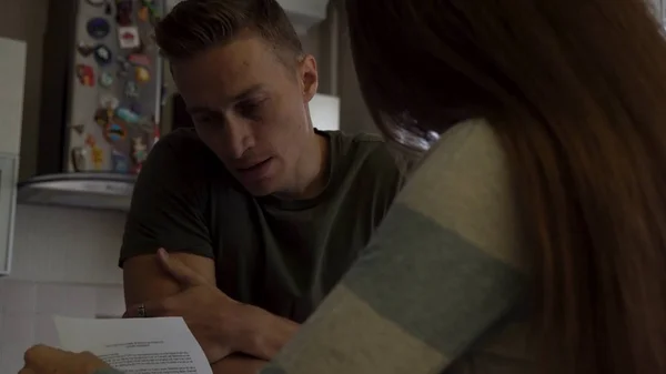 Worried couple reading together a letter standing near the table in the kitchen. Nervous man touching his mouth while his girlfriend opens a letter and begins to read.