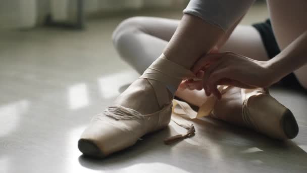 軽い木製の床にバレリーナの足のクローズ アップ。トウシューズのリボンを結ぶ少女. — ストック動画