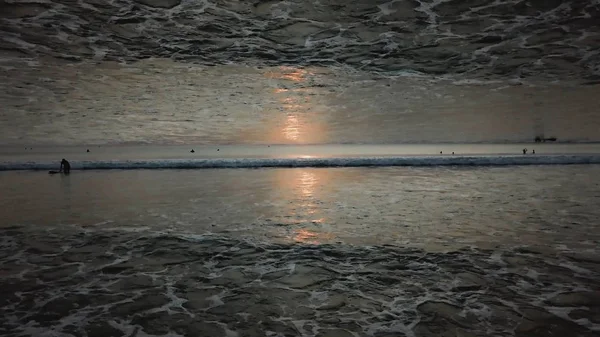 Atemberaubender Sonnenuntergangshimmel über dem Ozean mit Spiegelhorizonteffekt, Blick vom Ufer aus. endlose Wellen und Menschen im Wasser, Gründungsthema. — Stockfoto