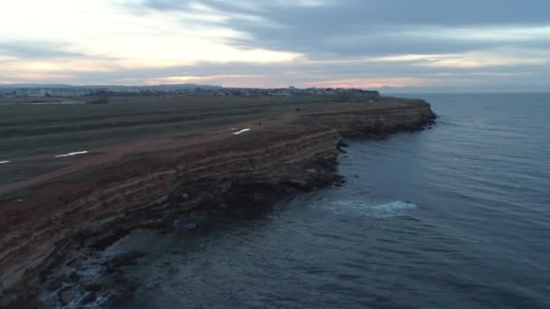 在乡村公路附近的海面前, 可以看到陡峭的悬崖, 在日落的天空中, 有移动的汽车和人们。拍摄。特写. — 图库视频影像