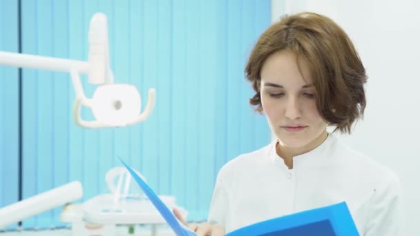 La doctora examina documentos con pruebas de pacientes. Medios. Mujer médico en bata blanca en el fondo de la sala de tratamiento lee el testimonio médico del paciente — Vídeos de Stock