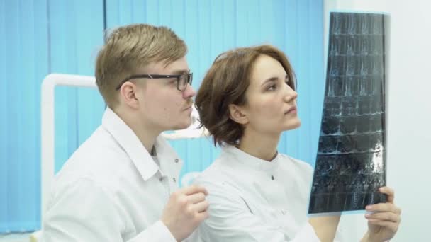 Two doctors are looking at x-rays. Media. Chief physician of male and female Intern look at x-rays MRI at bright light lamp — Stock Video