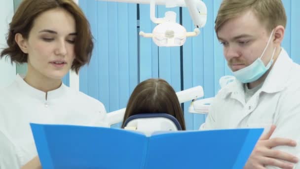 Dentysta męski i asystentka Dyskutować diagnozy pacjenta. Media. Kobieta i mężczyzna Dentyści twierdzą badania pacjent siedzi na krześle w tle — Wideo stockowe