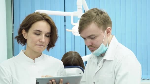 El dentista masculino y la asistente femenina discuten el diagnóstico de la paciente. Medios. Mujer y hombre dentistas argumentan pruebas de paciente sentado en la silla en el fondo — Vídeo de stock