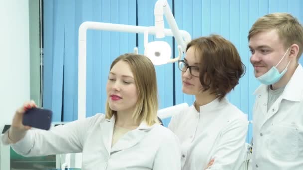 Jovens médicos fazem selfie. Mídia. Grupo de dentistas atraentes fazer selfies no fundo de equipamentos odontológicos durante o intervalo — Vídeo de Stock