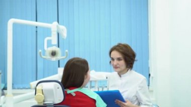 Diş hekimi tedavi hasta ile anlatılır. Medya. Başka bir dişçi istemciye konuşurken arka plan üzerinde çarpı işareti silah ile beyaz paltolu profesyonel diş hekimi duruyor