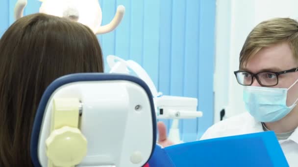 Mężczyzna dentysta konsultuje pacjenta w fotelu. Media. Dentysta, sprawdzanie dokumentów konsultuje się z pacjentem podczas badania stomatologicznego — Wideo stockowe