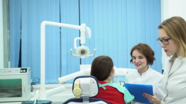 O dentista discute o tratamento com o paciente. Mídia. Dentista profissional de casaco branco fica com os braços cruzados no fundo de outro dentista falando com o cliente — Vídeo de Stock