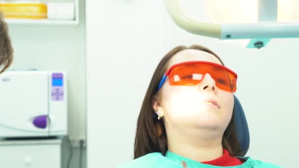 歯科用の椅子に座っていると、治療を待っているオレンジ色の保護メガネの若い女性患者のクローズ アップ。歯科医療. — ストック動画
