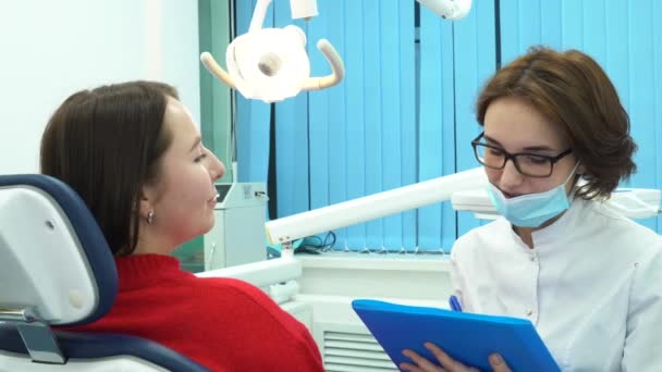 Primer plano de la paciente joven y su dentista con carpeta azul discutiendo el tratamiento en una consulta. Cuidado dental . — Vídeos de Stock