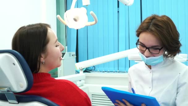 Nahaufnahme einer jungen Patientin und ihres Zahnarztes mit blauer Mappe, die die Behandlung auf einer Sprechstunde bespricht. Zahnpflege. — Stockvideo
