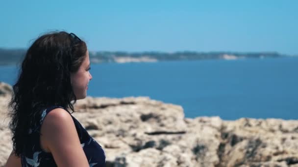 Genç kız bir uçurumun kenarında duruyor ve denizde görünüyor. Güzel kız deniz cliff bir manzaraya sahip. Kız uçurumun kenarına okyanus manzarası hoşlanır — Stok video