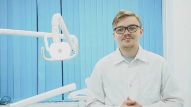 Diş hekimi diş ekipman arka plan üzerinde gözlük takıyor. Medya. Genç diş hekimi gözlük koyar ve gülümseme diş Office ile selamlıyor