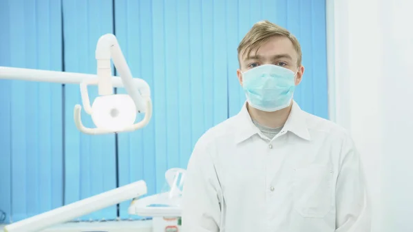 Docteur en masque et manteau blanc sur le fond de la salle de traitement. Les médias. Attrayant médecin masqué est prêt à commencer à traiter le patient — Photo