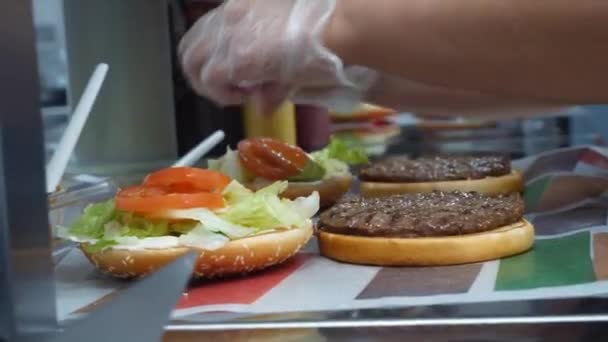 Primer plano sobre la preparación de hamburguesas clásicas. Una trampa. Chef agrega ensalada y tomates a la chuleta frita en un bollo de hamburguesa . — Vídeos de Stock