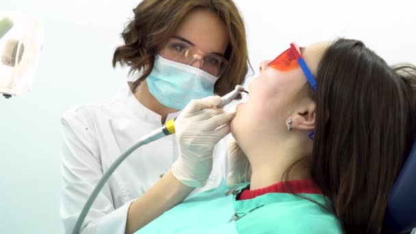 Jovem sentada na cadeira dentária no exame médico no consultório odontológico. Mídia. Vista lateral da bela dentista feminina que trata os dentes dos pacientes . — Vídeo de Stock