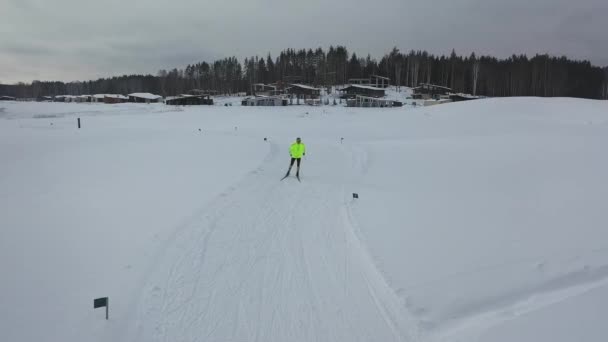 Vista aérea do homem em roupas amarelas em uma pista de esqui perto da floresta e pousadas de esqui no inverno contra o céu cinzento da noite. Filmagem. Esportes e recreação — Vídeo de Stock