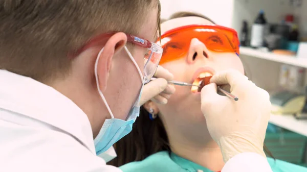 Professionele tandarts werken met patiënt in moderne kliniek, geneeskunde concept. Media. Jonge patiënt in beschermende bril terwijl tanden behandelen proces. — Stockfoto