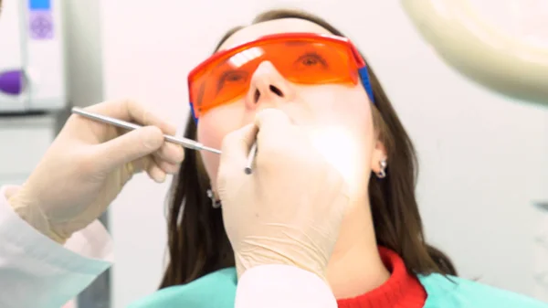 Vrouwelijke patiënt met een arts proberen om haar te onderzoeken in het kantoor van een tandarts, tandheelkundige zorg concept. Media. Een jong meisje in beschermende bril zittend in de stoel bij de tandarts bezoek. — Stockfoto