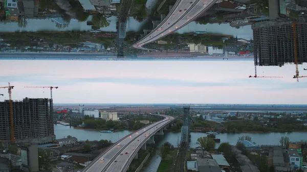 Vista de la mañana temprano de la ciudad con puente de río, edificio inacabado y muchos árboles verdes, efecto espejo horizonte. Medios. Mañana gris en la ciudad con el río turquesa, barcos y el puente . —  Fotos de Stock