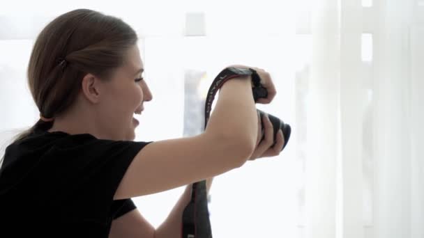 Fotógrafo olha para a câmera tirando fotos e sorri para o fundo branco. Mídia. Vista lateral da jovem fotógrafa profissional filmando com sorriso na câmera — Vídeo de Stock