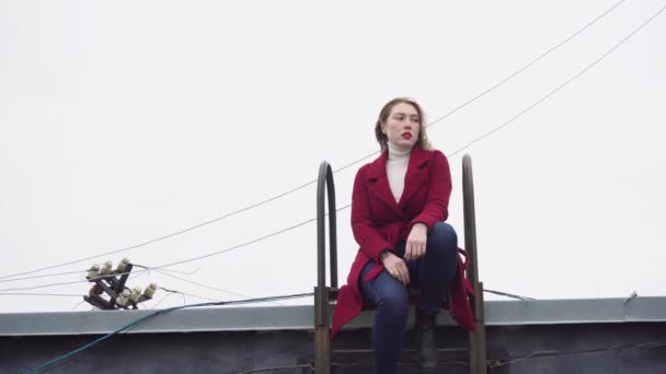 Modèle assis sur le toit en hiver. Les médias. attrayant jeune femme modèle posant assis sur le toit avec échelle sur fond de fils suspendus et ciel nuageux — Video