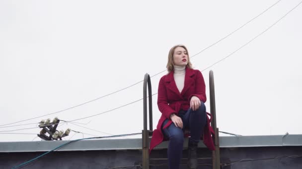 Model kışın çatıda oturuyor. Medya. Çekici genç kadın model asılı teller ve bulutlu gökyüzü arka planda merdiven ile çatıda oturan poz — Stok video