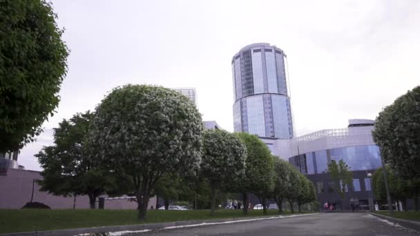 Beautiful spring cityscape with modern buildings standing behing apple bushes. Stock footage. Blossoming apple trees against the background of glass facade buildings. — Stock Video