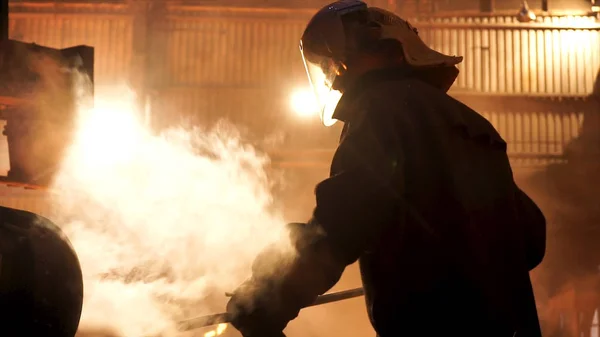 Steel worker removing slag from the electric induction crucible melting furnace at the metallurgical plant, hard work conceprt. Stock footage. Man in protective mask and uniform working with a poker.