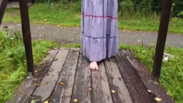 Close-up of pretty young woman with red hair in long skirt walking on a wooden bridge in park. Country walks. — Stock Video