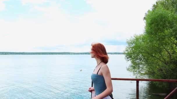 Mavi gömlekli kırmızı saçlı ve nehir kenarındaki bir iskelede duran açık etekli genç bir kadının yakın çekim. Ülke yürüyüşleri. — Stok video